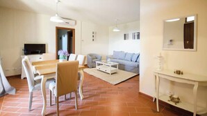 A living room with a sofa, coffee table, and TV. There is a dining area with a table and chairs. The room has a tiled floor and white walls.