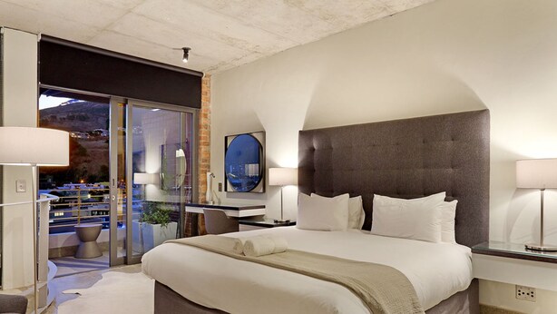 Ein modernes Hotelzimmer mit einem großen Bett, einer Glastür, die auf einen Balkon führt, und Blick auf die Stadt.