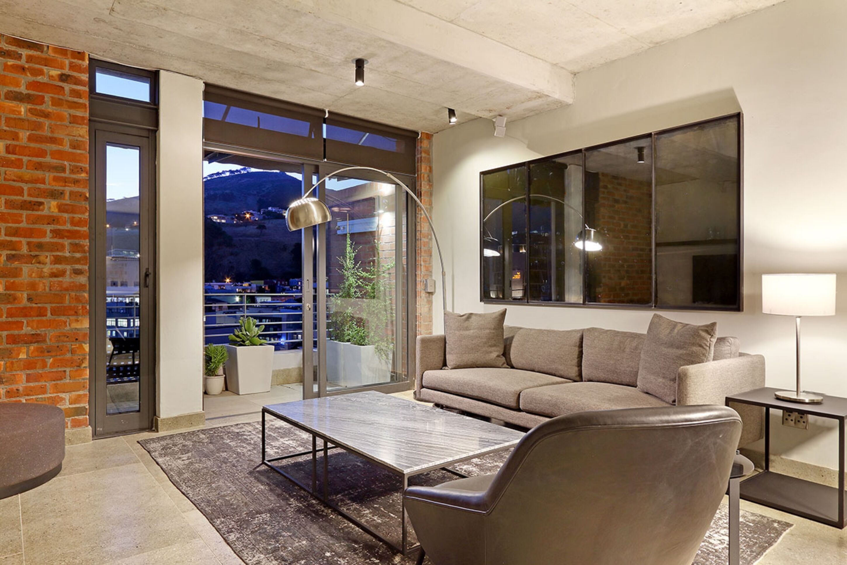Un salon moderne avec un canapé, une table basse et une lampe. Une grande fenêtre offre une vue sur le paysage urbain et les montagnes.