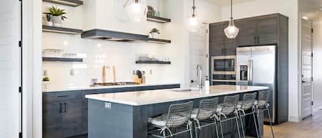 Una cocina moderna con una isla central, taburetes de bar, electrodomésticos integrados e iluminación colgante.