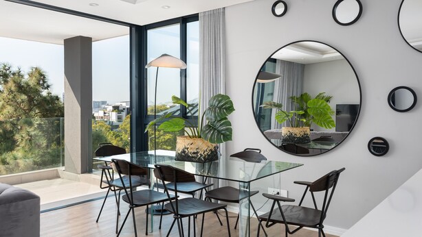 Un comedor moderno con una mesa y sillas de vidrio, ventanas amplias y plantas en macetas.