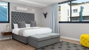Un dormitorio moderno con un cabecero capitonado, un ventanal con vistas a edificios, una decoración de pared con forma de cabeza de toro y un otomano amarillo brillante.