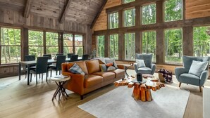A spacious living area with a high ceiling, large windows, a wooden interior, a leather sofa, a coffee table, and a dining area with chairs and a table.