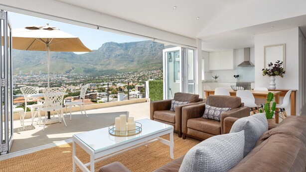 Ein modernes Wohnzimmer mit einem Glastisch, braunen Sofas und Blick auf eine Stadt und Berge.