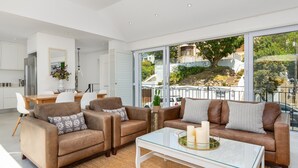 Ein modernes Wohnzimmer mit zwei braunen Ledersofas, einem Glastisch und Blick auf eine Wohngegend durch große Fenster.