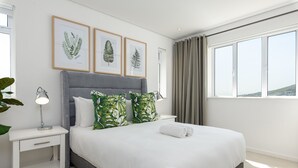 Ein ordentlich bezogenes Bett mit grauem Kopfteil, weißer Bettwäsche und grün gemusterten Kissen. An der Wand hängen gerahmte Pflanzenbilder, eine Nachttischlampe und ein Fenster mit Vorhängen.
