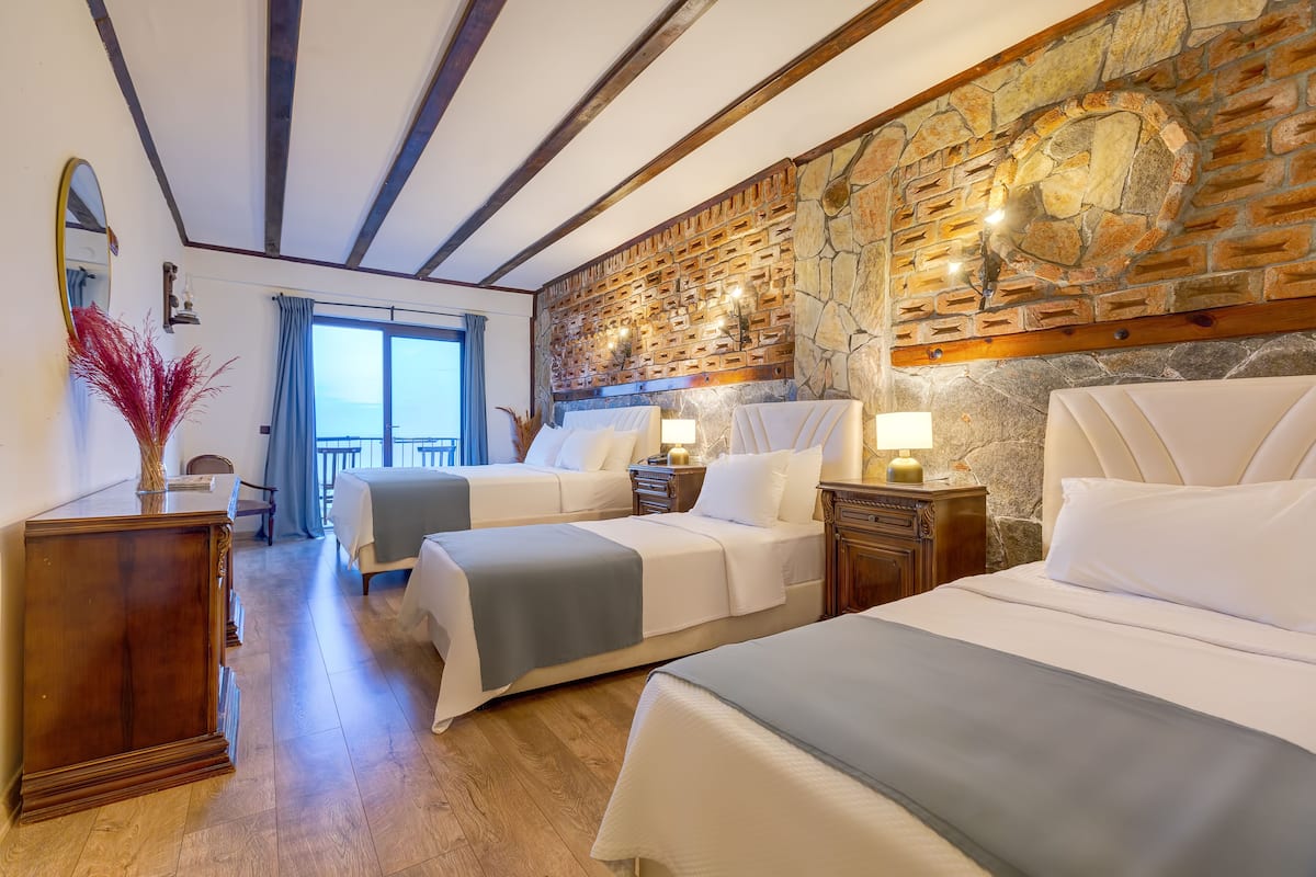 A hotel room with two beds, a stone wall, wooden beams, a large window, and a view of the sea.