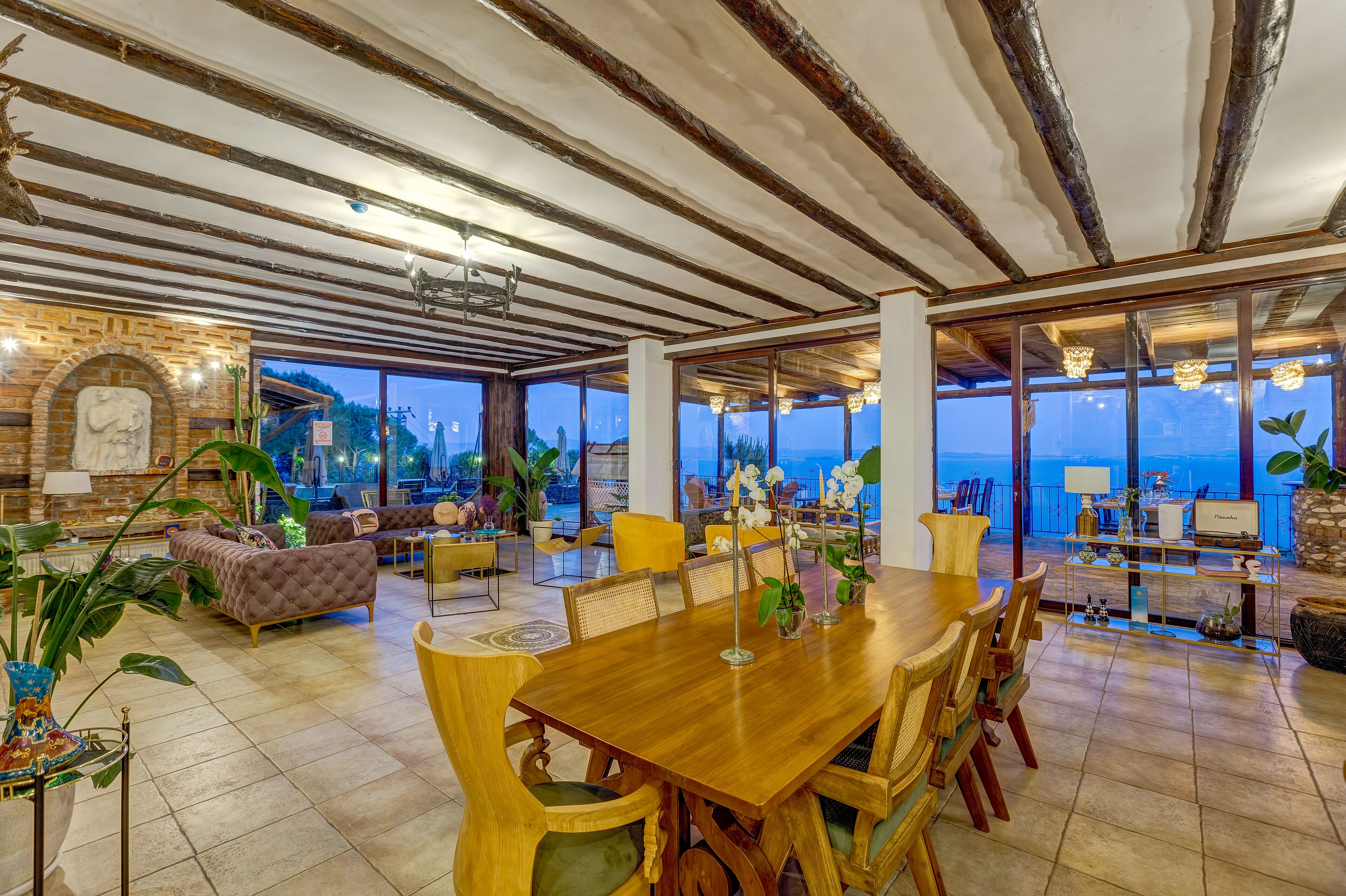 A spacious living area with a wooden dining table, comfortable seating, and large windows offering a view of the sea.