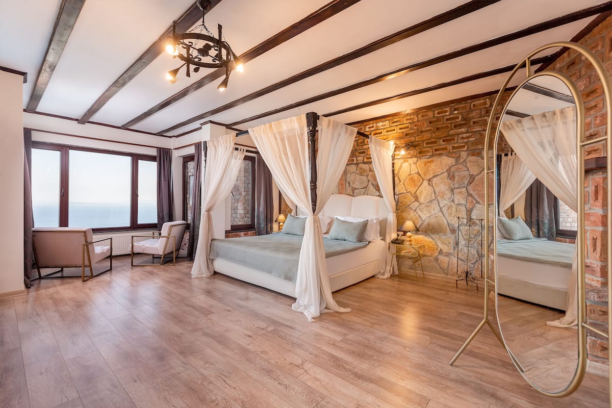 A bedroom with a large bed, a sitting area with a chair, a small table, and a large mirror.
