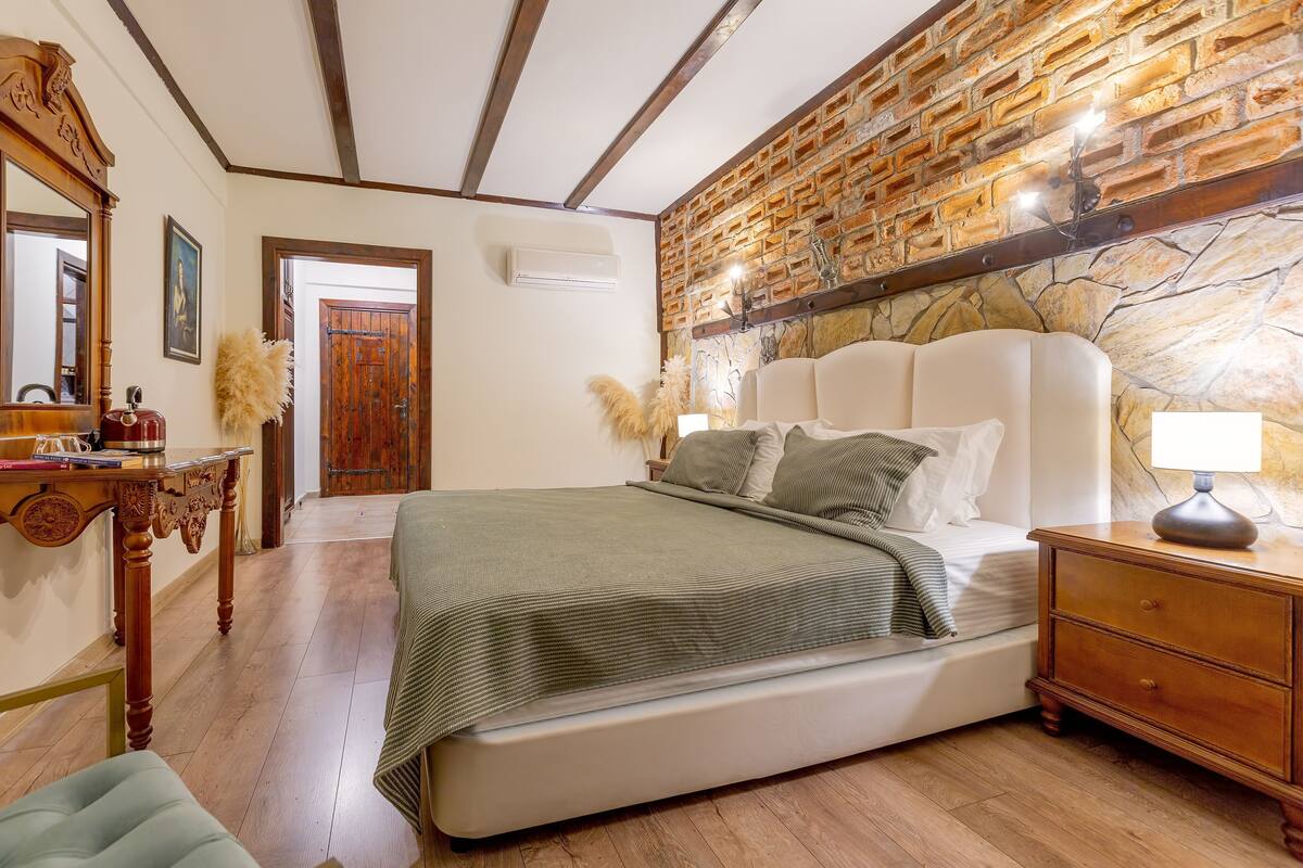A bedroom with a brick wall, a wooden headboard, a bedside table with a lamp, and a bed with a green blanket.
