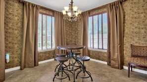 A dining area with a round table and four stools, flanked by two windows with blinds, a chandelier, and patterned wallpaper. - Tuscan Getaway in the Heart of Gilbert! (Gilbert)