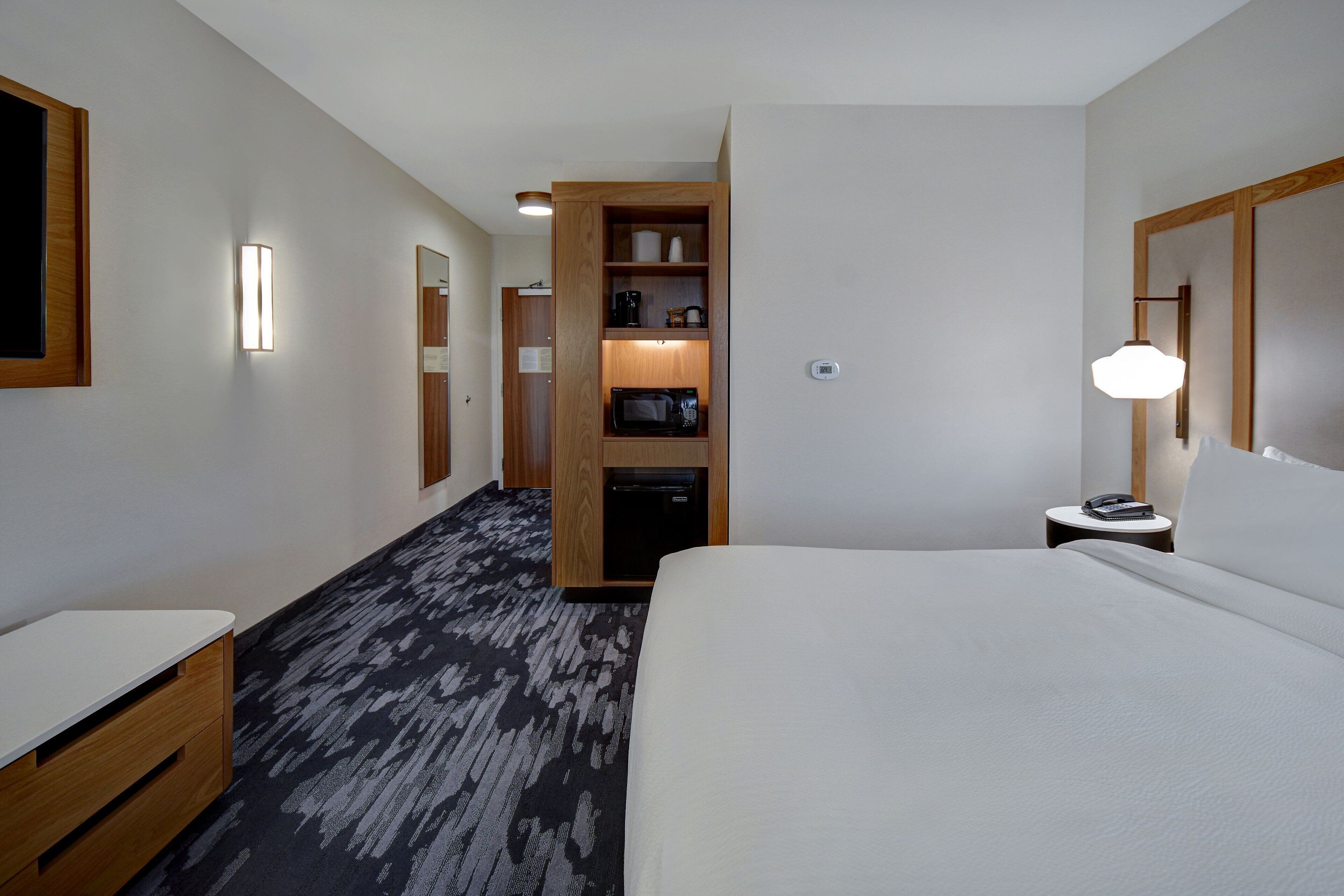 Een hotelkamer met een bed, een bureau, een kledingkast, een magnetron en een nachtkastje met een lamp.