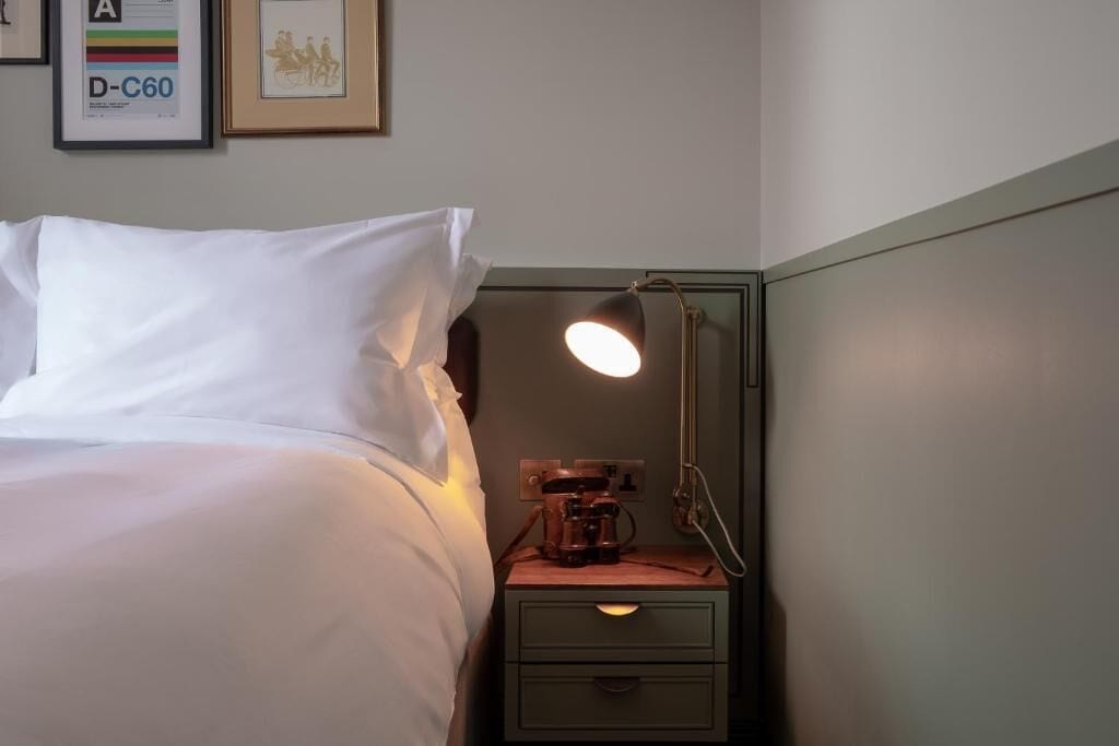 Un lit bien fait, avec du linge de lit blanc, une table de chevet avec une lampe et un tableau encadré au mur.