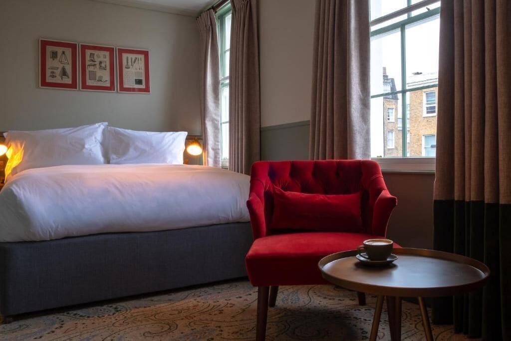 A hotel room with a bed, a red armchair, a small table with a cup, and a window with curtains.