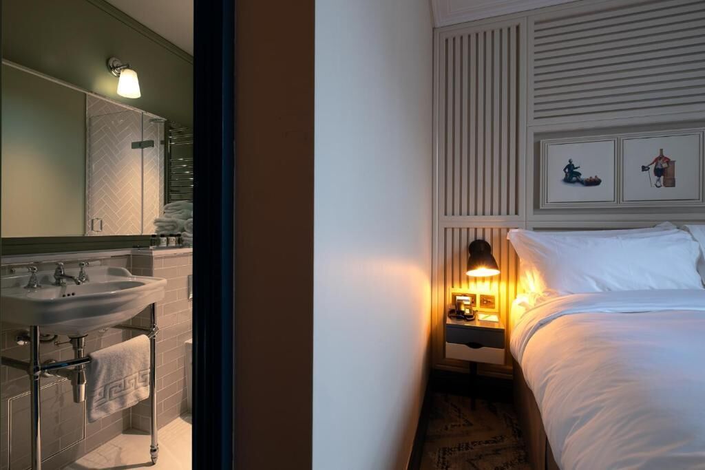A hotel room with a bathroom, a bed, a nightstand with a lamp, and artwork on the wall.