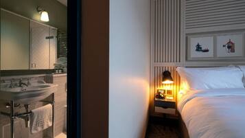A hotel room with a bathroom, a bed, a nightstand with a lamp, and artwork on the wall.