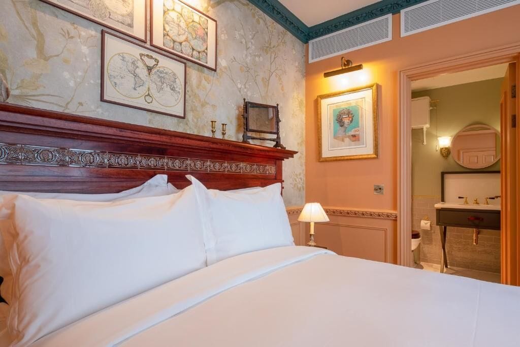 A hotel room with a large bed, a wooden headboard, framed artwork on the walls, a bedside lamp, and a bathroom visible through an open door.