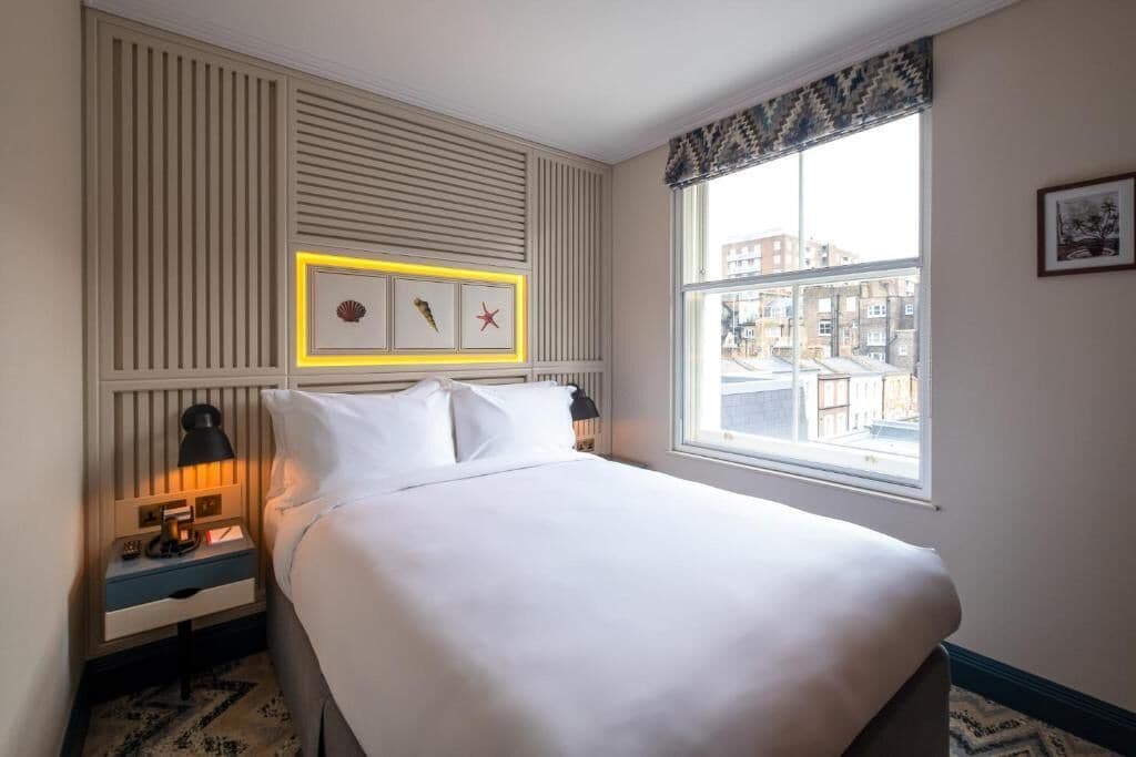 A hotel room with a bed, bedside table, lamp, window with view, and wall decor.