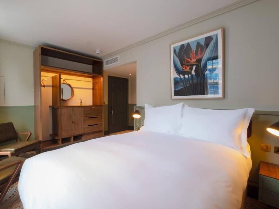 A hotel room with a large bed, a wooden dresser, a chair, a desk, and a painting on the wall.