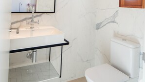 A modern bathroom with a marble wall, a white sink, a toilet, and a wooden-framed window.
