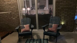 Twee Adirondack stoelen met kussens met Amerikaanse vlag, een groen-wit gestreepte mat en een bakstenen muur.