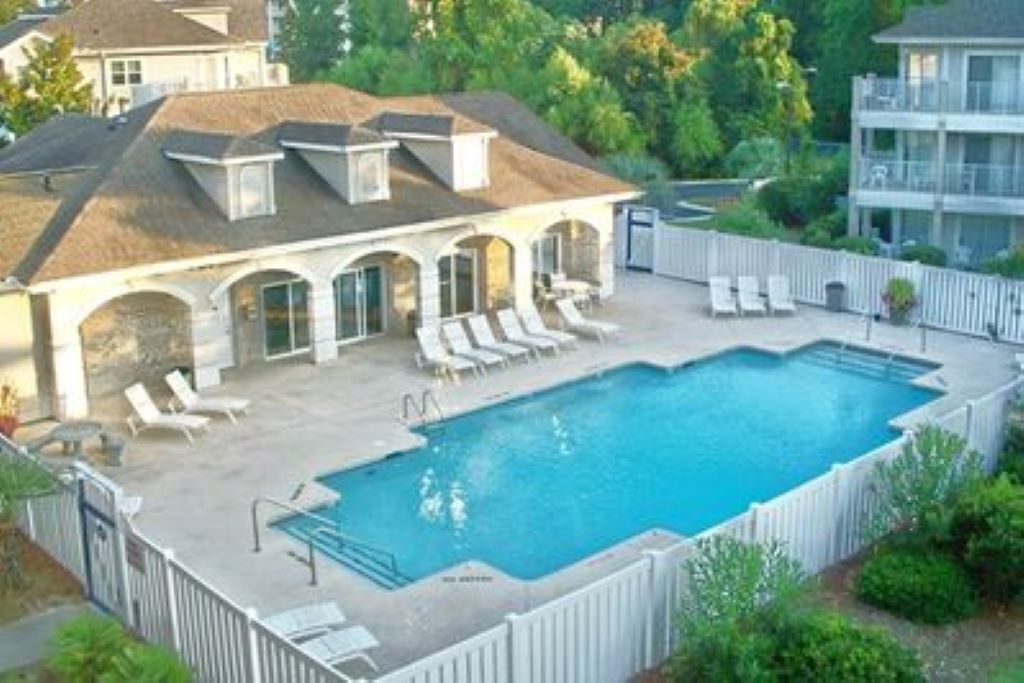 Ein Schwimmbecken mit Liegestühlen, eine umzäunte Terrasse und ein Gebäude mit Satteldach und Rundbogenfenstern.