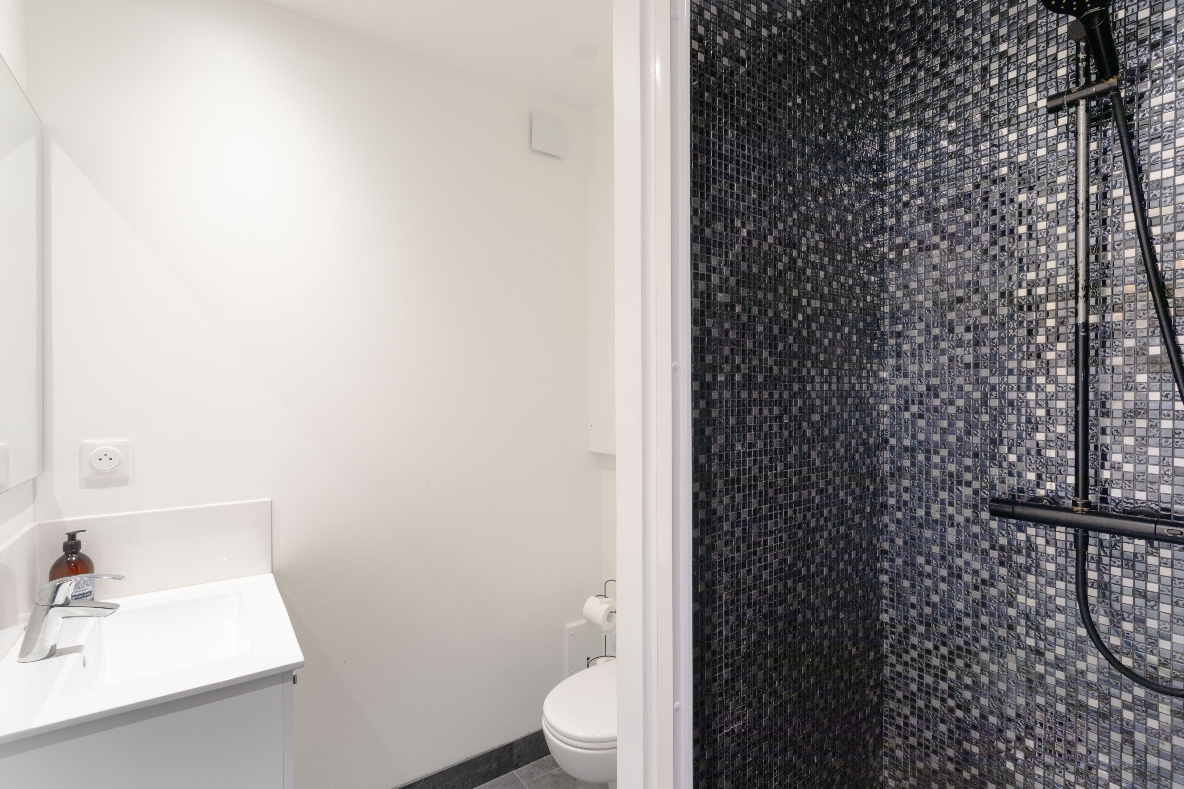Un baño con lavamanos blanco, inodoro y ducha con pared de mosaicos.