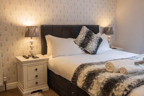 A neatly made bed with white linens and a fluffy blanket, flanked by two bedside lamps, a nightstand with a remote control, and a wall with a subtle pattern. - Ironmasters House (Telford)
