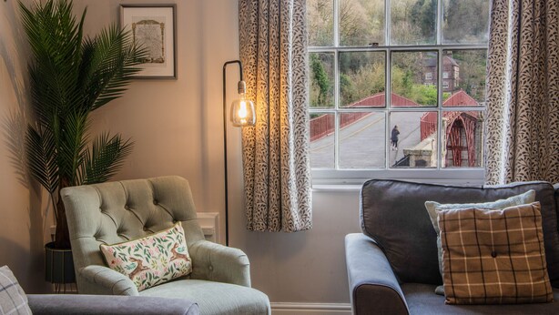 A room with a large window, a chair with a cushion, a sofa with a cushion, a potted plant, and a wall-mounted light fixture. - Ironmasters House (Telford)