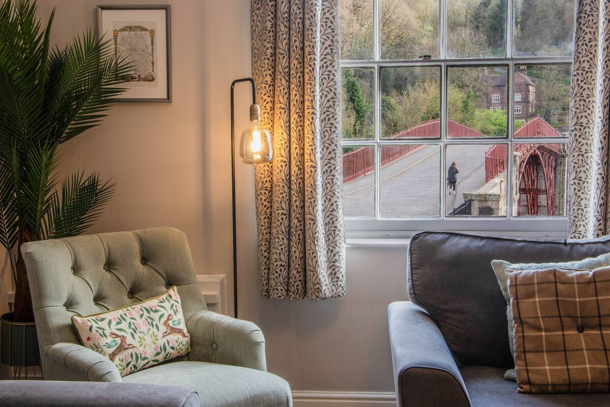 A living room with a tufted armchair, a sofa, a large potted plant, and a window with patterned curtains.