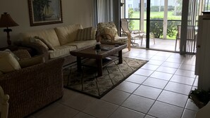 A living room with a sofa, armchair, and coffee table. There is a painting on the wall and a view of a garden through the glass door. - 2-Bedroom Condo Steps to Beach, Pools, and Golf (Fort Pierce)
