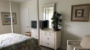 A bedroom with a bed, a dresser, a chair, a television, a mirror, and a potted plant. - 2-Bedroom Condo Steps to Beach, Pools, and Golf (Fort Pierce)