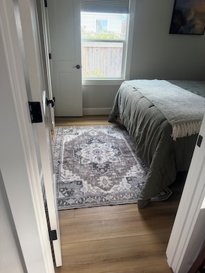 A bedroom with a bed, a patterned rug, wooden flooring, a door, and a window with a view of the outside.