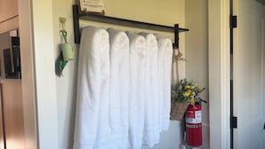Un baño con un perchero que sostiene toallas blancas, un certificado enmarcado, un extintor y una mesita con una botella.