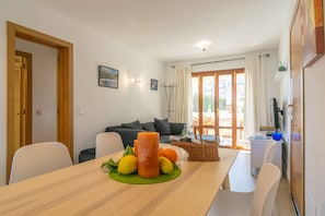 A dining area with a wooden table, white chairs, and a basket. There is a sofa and a window with curtains in the background.