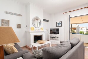 Uma sala de estar com sofá cinzento, lareira branca, televisão e varanda com vista.