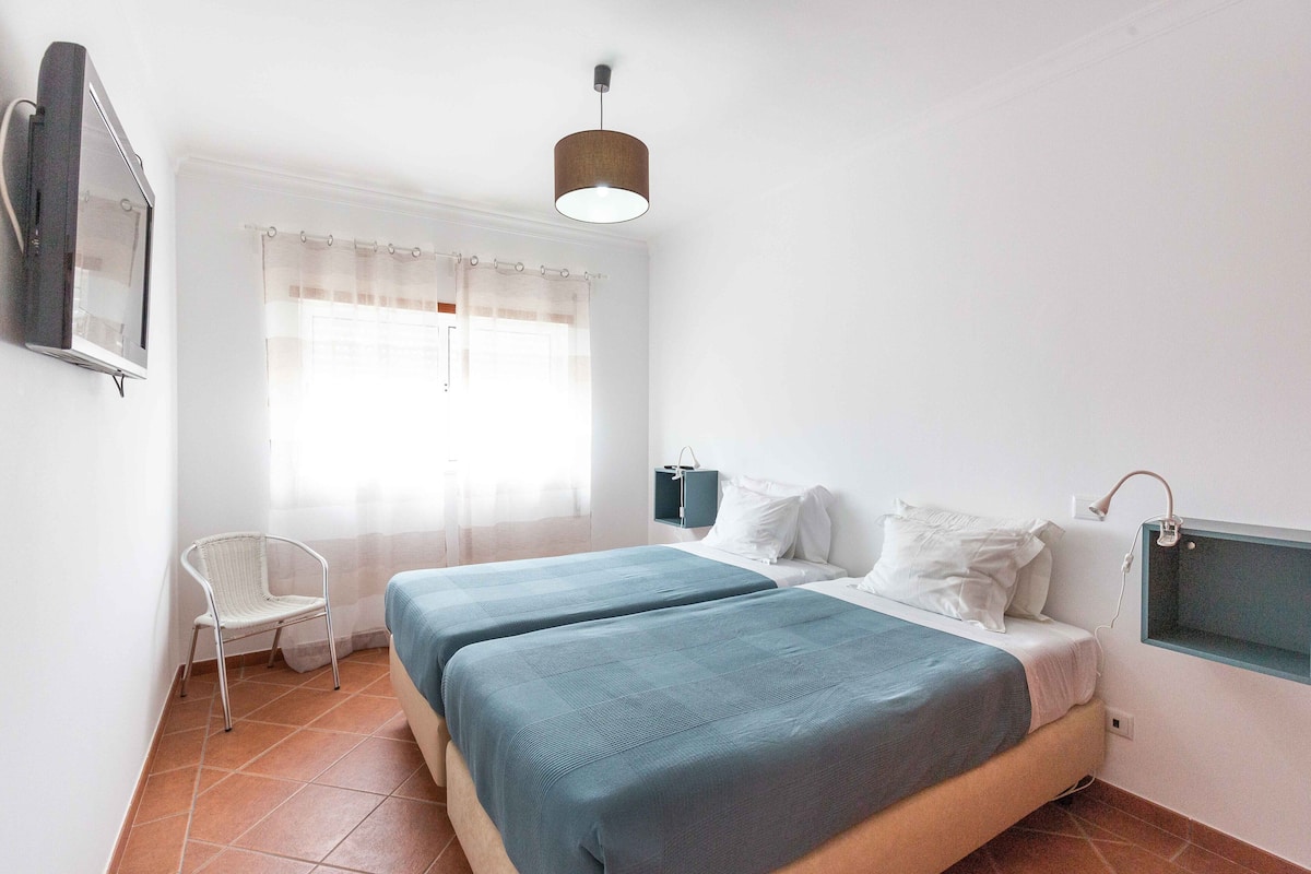 A bedroom with two beds, a chair, a TV, and a window with curtains.