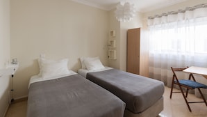 Ein Hotelzimmer mit zwei Einzelbetten, einem kleinen Tisch, einem Stuhl und einem Fenster mit durchsichtigen Vorhängen.