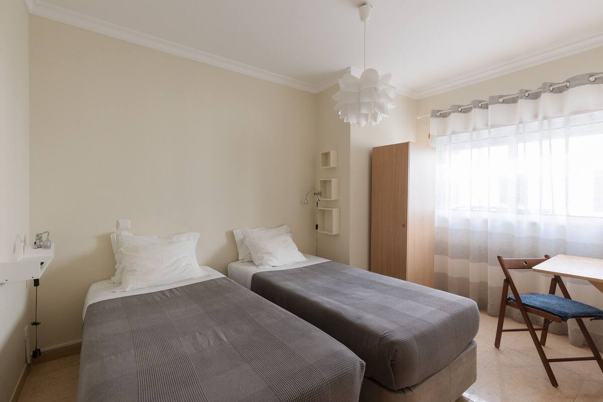 A hotel room with two single beds, a small table, a chair, and a window with sheer curtains.