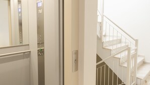 Ein Aufzug mit Bedienfeld und Glastür, der zu einer Treppe mit weißer Geländer und Marmorboden führt.