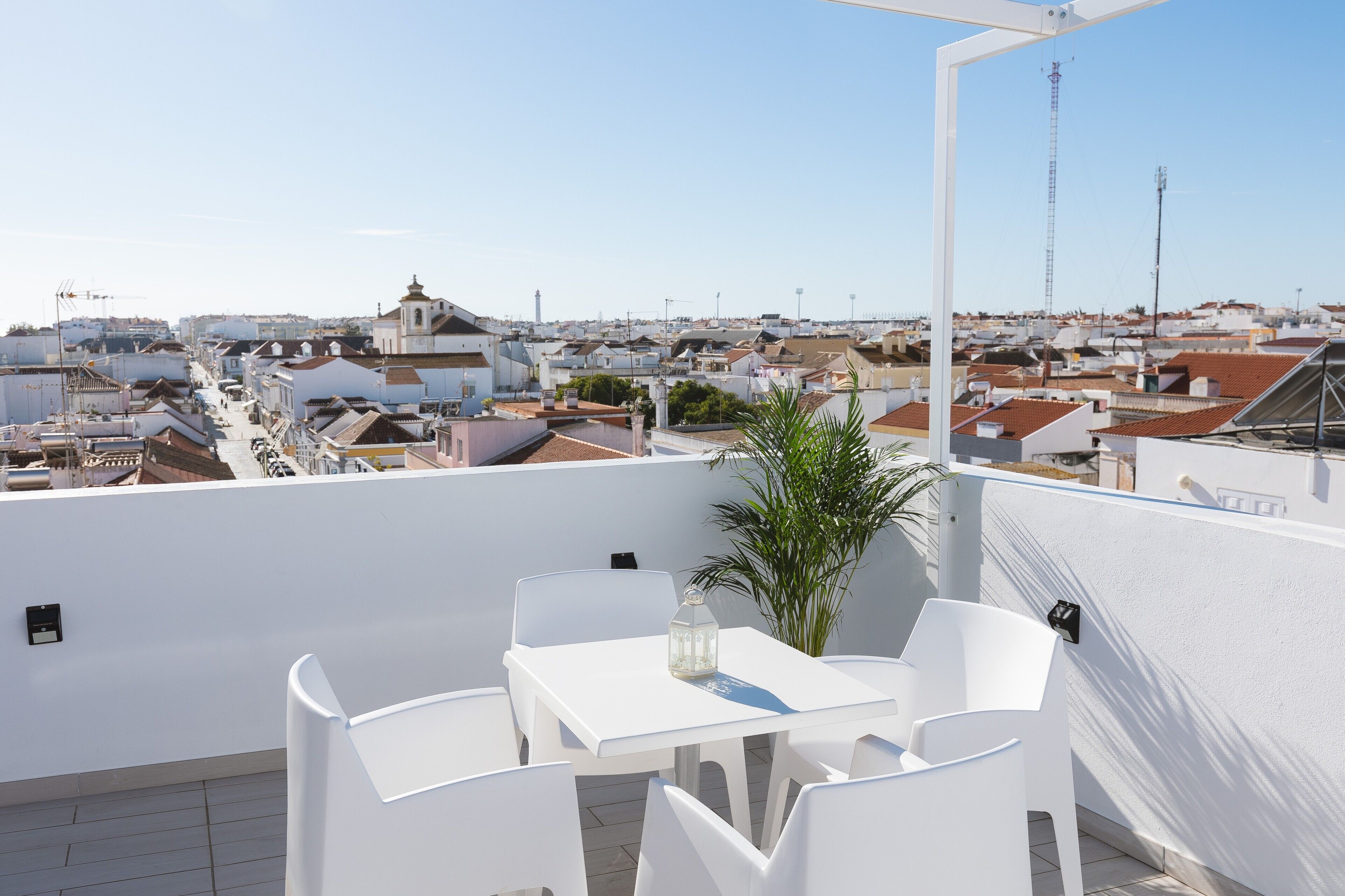 Eine Dachterrasse mit weißem Tisch und Stühlen, einer Topfpflanze und freiem Blick auf die Stadtlandschaft.