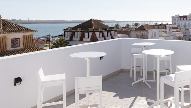 Eine Dachterrasse mit weißen Tischen und Stühlen, mit Blick auf ein Gewässer und Gebäude.