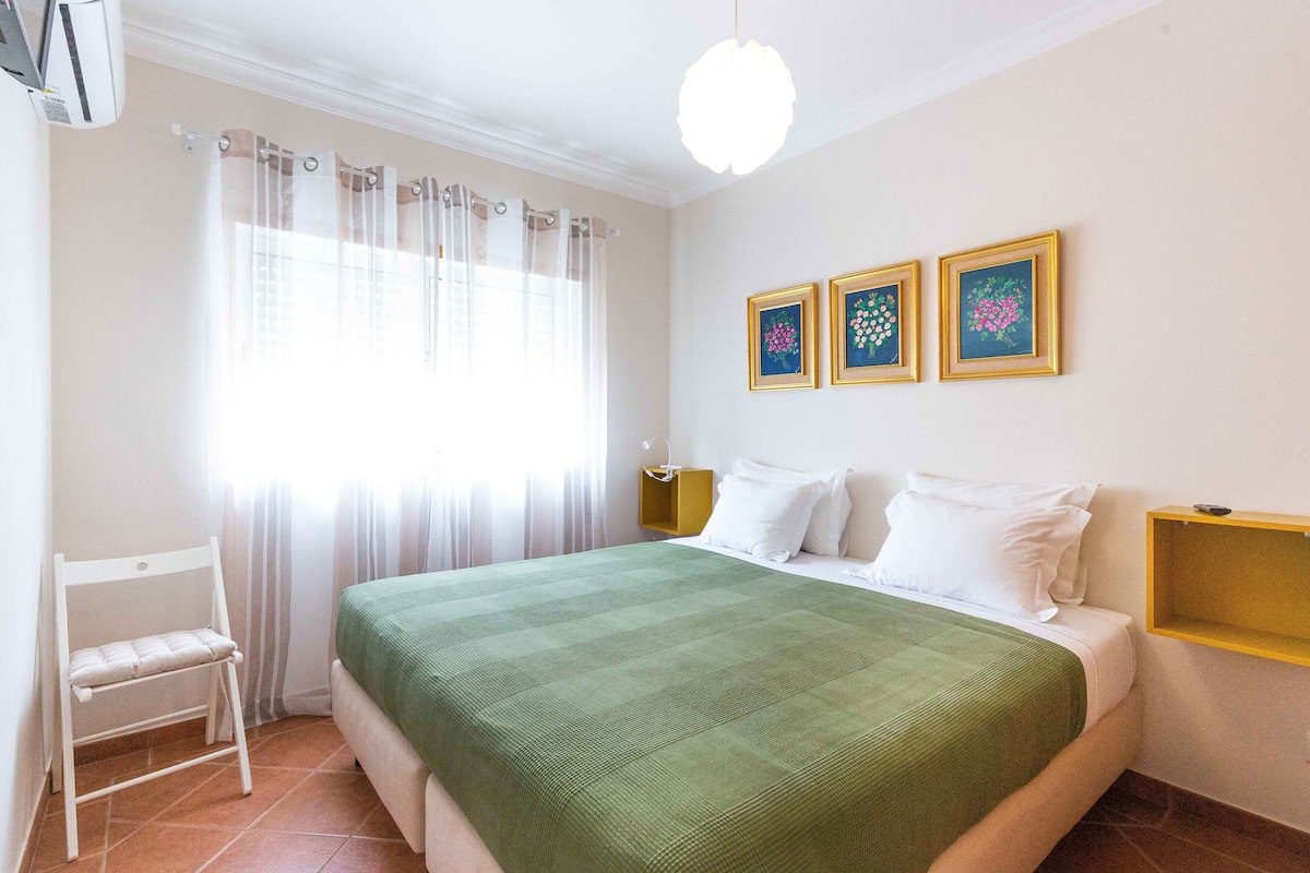 A neatly arranged bedroom with a bed, a chair, a nightstand, and a wall-mounted shelf.