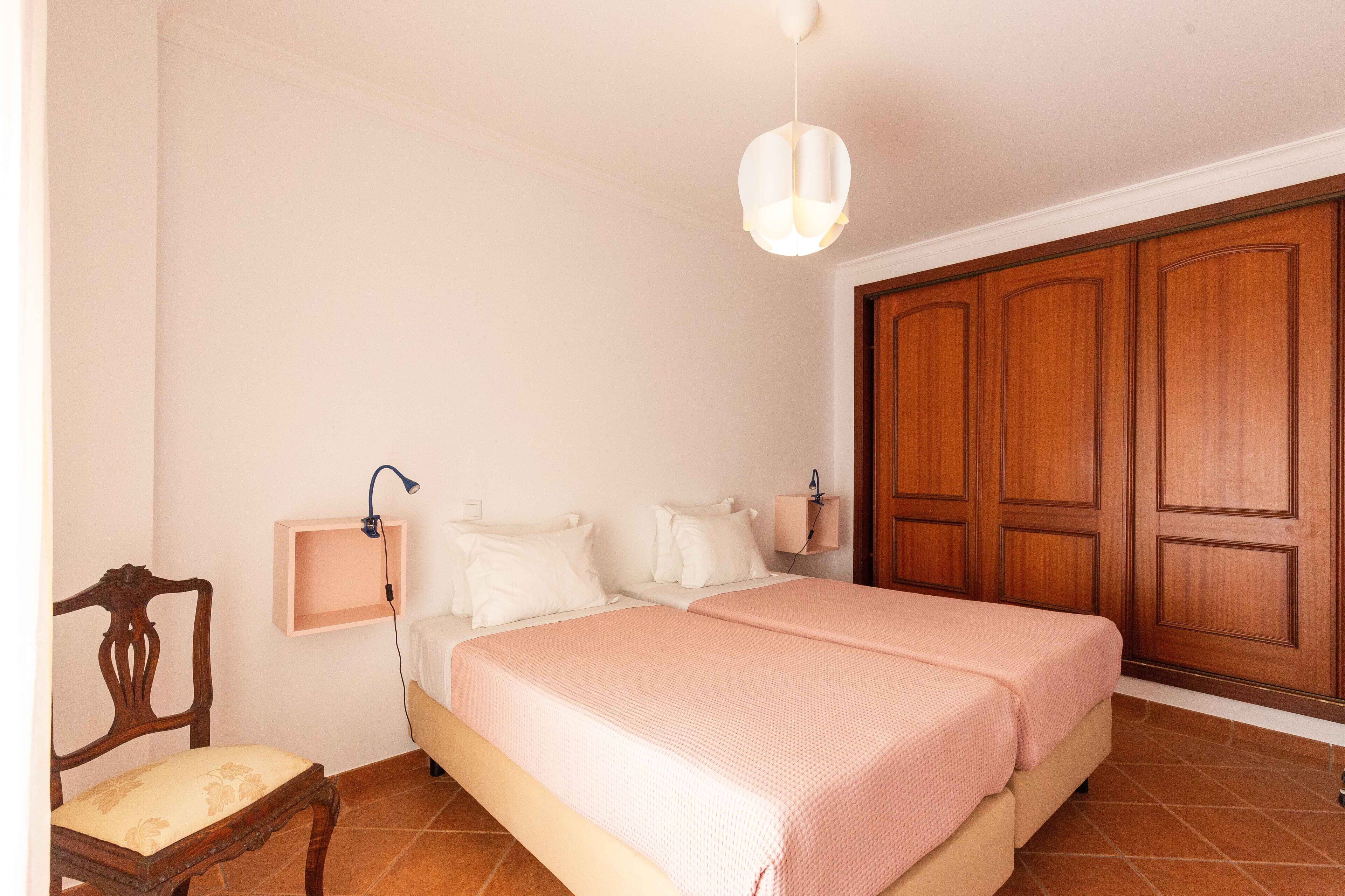 A bedroom with a double bed, a wooden wardrobe, a bedside table with a lamp, and a chair.
