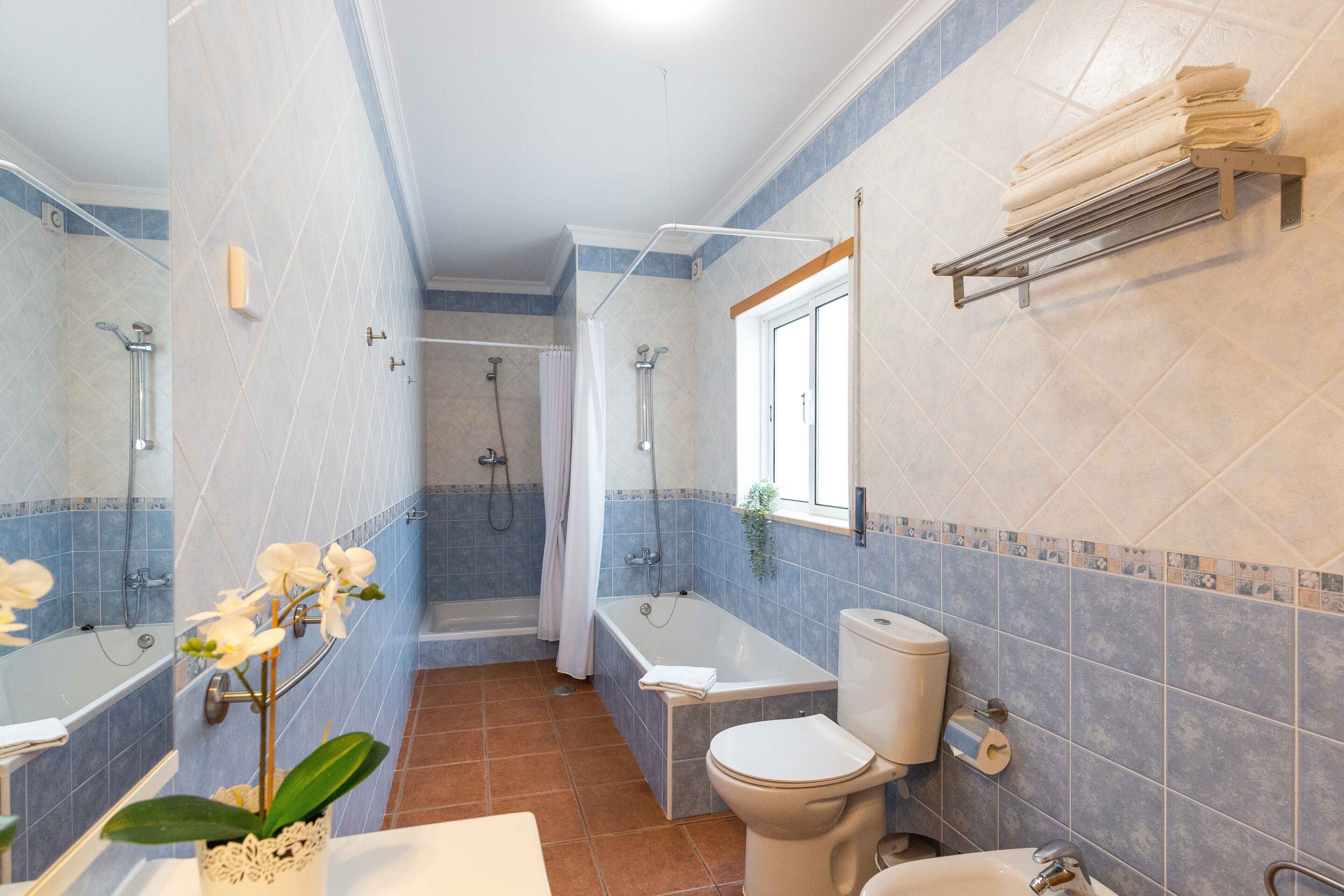 Un baño con bañera, inodoro y lavabo, con azulejos azules y beige, una planta y un perchero para toallas.
