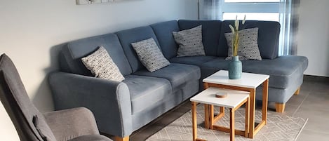 A modern living room with a grey sofa, a wooden coffee table, a patterned rug, and a blue knitted pouf.