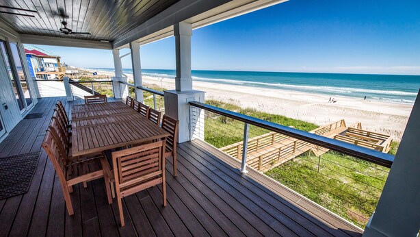 Une terrasse en bois avec une table et des chaises, donnant sur une plage et offrant une vue dégagée sur l’océan et le rivage sablonneux.