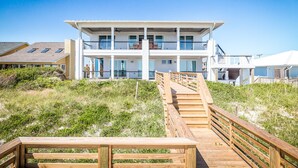 Une maison à plusieurs étages située en bord de mer, avec une terrasse en bois menant à l’entrée.
