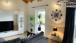 Ein modernes Wohnzimmer mit grauem Sofa, Flachbildfernseher, Topfpflanzen, einer Wanduhr und einem kleinen Tisch mit Lampe.