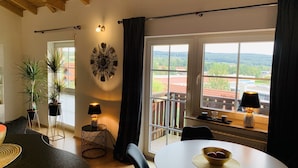 Ein modernes Wohnzimmer mit rundem Tisch, zwei Blumentöpfen, einer Wanduhr und Blick auf eine Stadt durch große Fenster.